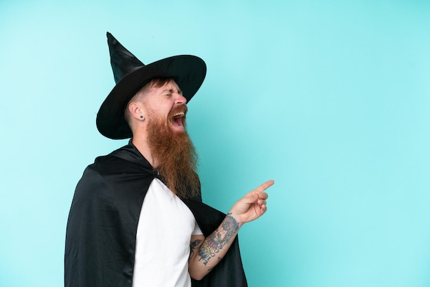 Young wizard in halloween isolated on blue background pointing finger to the side and presenting a product