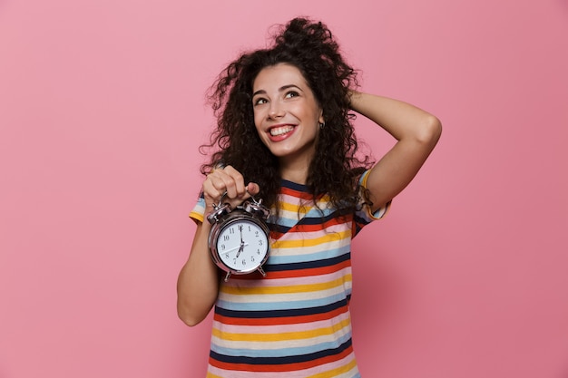 young woman 20s with curly hair holding alarm clock isolated on pink