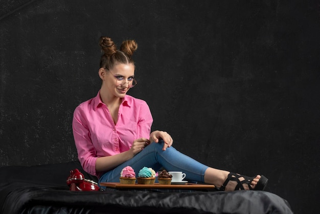 Young woman admires cupcakes with multicolored cream Beautiful girl with glasses sits on black background