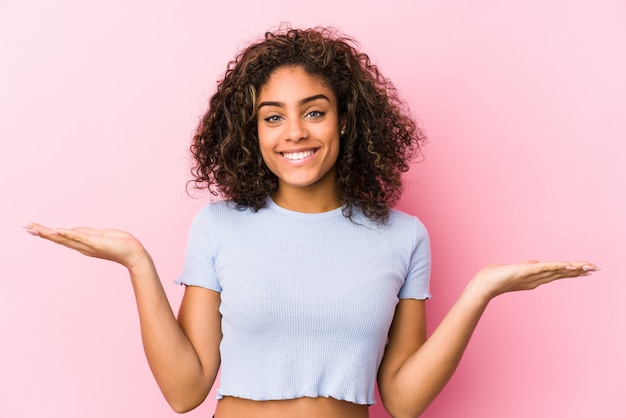 Young woman against a pink makes scale with arms, feels happy and confident.