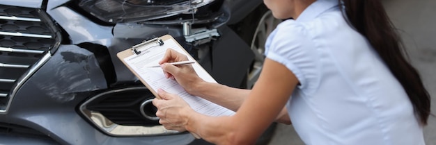 写真 壊れた車の近くに座って、ドキュメントに情報を書き込む若い女性エージェント自動車保険