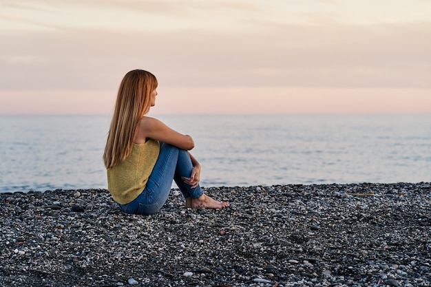 写真 日没時にビーチの砂の上に一人で座っている若い女性。リラクゼーションと瞑想の概念