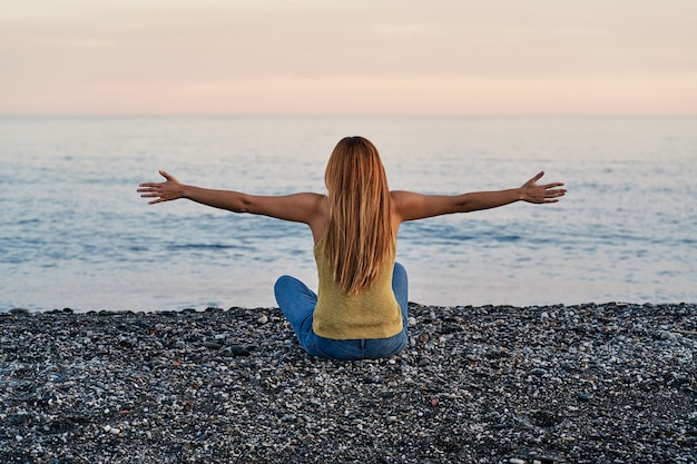 젊은 여자 혼자 석양 해변 모래에 두 팔을 벌려 앉아. 휴식, 명상 및 자유의 개념.