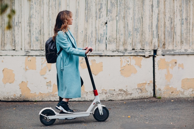 Pendolare alternativo della giovane donna con il motorino elettrico in cappotto blu alla città