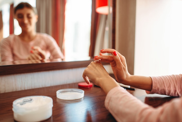 Young woman applies hand cream to skin at the mirror in the bedroom. Morning skincare procedure