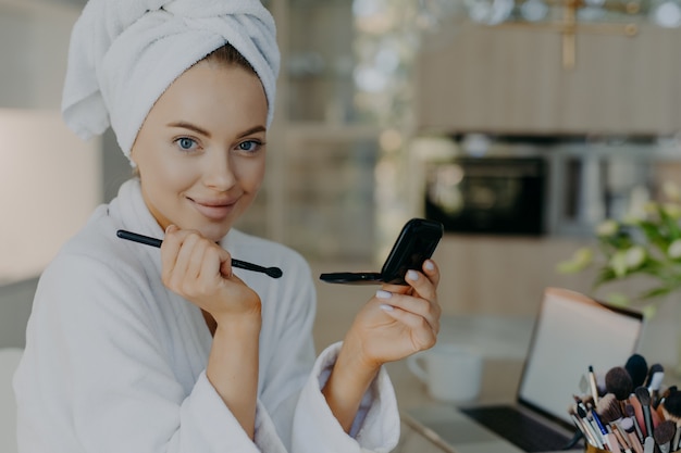 young woman applies makeup cosmetics with brush in front of mirror