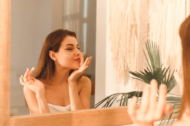 Молодая женщина наносит увлажняющий крем на лицо в солнечной ванне Ежедневная рутинная концепция увлажнения и ухода за кожей