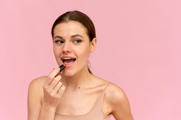 Photo a young woman applies nude lipstick on her lips on a pink background. cosmetics concept.