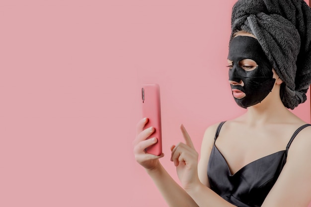 若い女性は、黒の化粧品ファブリックフェイシャルマスクと電話を手に適用します。チャコール、スパエステ、スキンケア、美容のあるフェイスピーリングマスク。閉じる