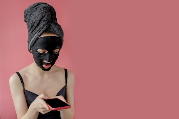 Young woman apply black cosmetic fabric facial mask and phone\
in hands on pink background. face peeling mask with charcoal, spa\
beauty treatment, skincare, cosmetology. close up