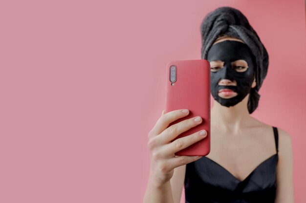 Young woman apply black cosmetic fabric facial mask and phone in hands on pink background Face peeling mask with charcoal spa beauty treatment skincare cosmetology Close up