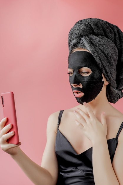 Young woman apply black cosmetic fabric facial mask and phone in hands on pink background. Face peeling mask with charcoal, spa beauty treatment, skincare, cosmetology. Close up