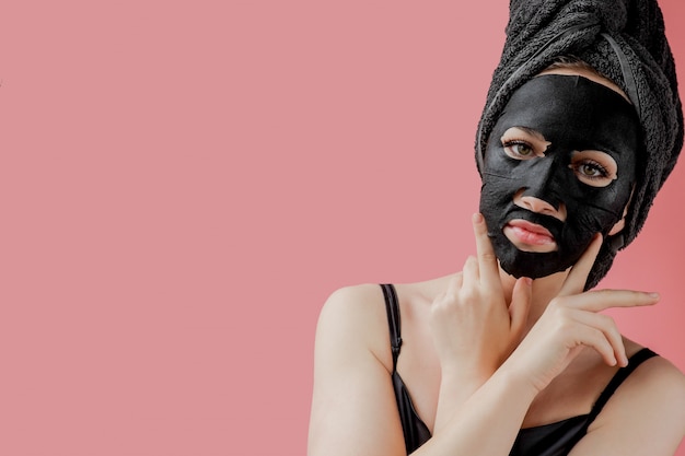 Young woman apply black cosmetic fabric facial mask on pink\
background. face peeling mask with charcoal, spa beauty treatment,\
skincare, cosmetology. close up