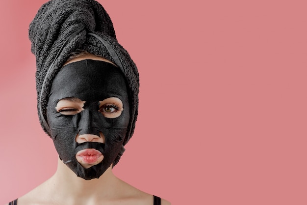 Young woman apply black cosmetic fabric facial mask on pink\
background. face peeling mask with charcoal, spa beauty treatment,\
skincare, cosmetology. close up