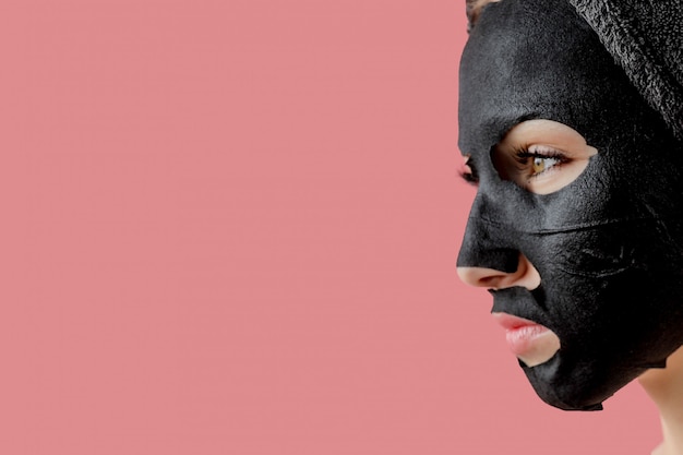 Young woman apply black cosmetic fabric facial mask on pink\
background. face peeling mask with charcoal, spa beauty treatment,\
skincare, cosmetology. close up