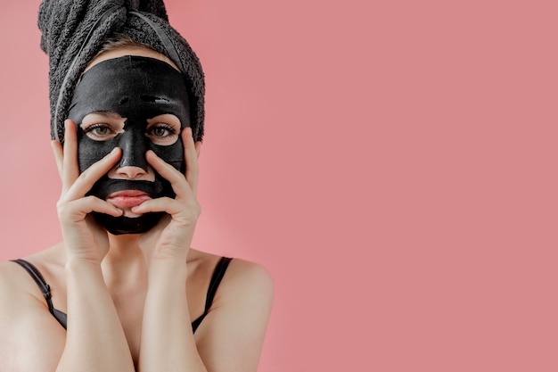 Young woman apply black cosmetic fabric facial mask on pink background. Face peeling mask with charcoal, spa beauty treatment, skincare, cosmetology. Close up