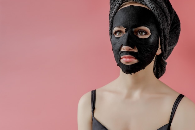 Young woman apply black cosmetic fabric facial mask on pink\
background. face peeling mask with charcoal, spa beauty treatment,\
skincare, cosmetology. close up