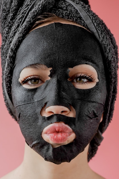 La giovane donna applica la maschera facciale nera del tessuto cosmetico sulla parete rosa. maschera peeling viso con carbone, trattamenti di bellezza spa, cura della pelle, cosmetologia. avvicinamento