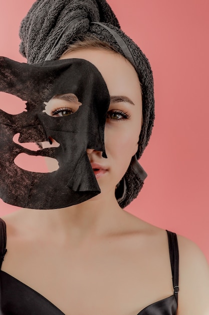 Young woman apply black cosmetic fabric facial mask on pink
wall. face peeling mask with charcoal, spa beauty treatment,
skincare, cosmetology. close up