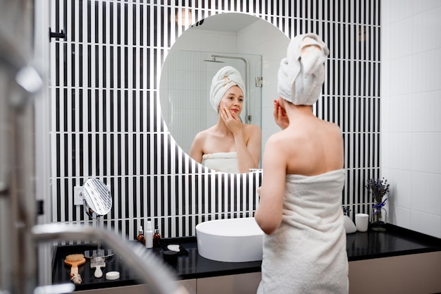 Young woman applying antiwrinkle cream standing behind mirror in home bathroom Cosmetology and beauty procedure Skin care after cleansing