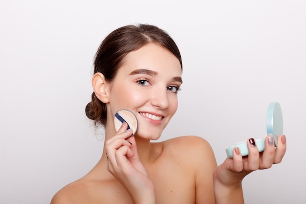 Young woman applying blusher on her face with powder puff isolated on white wall