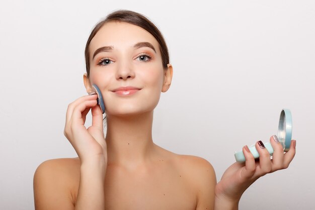 Young woman applying blusher on her face with powder puff isolated on white wall