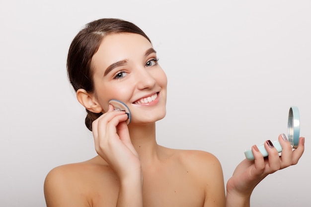 Young woman applying blusher on her face with powder puff isolated on white wall