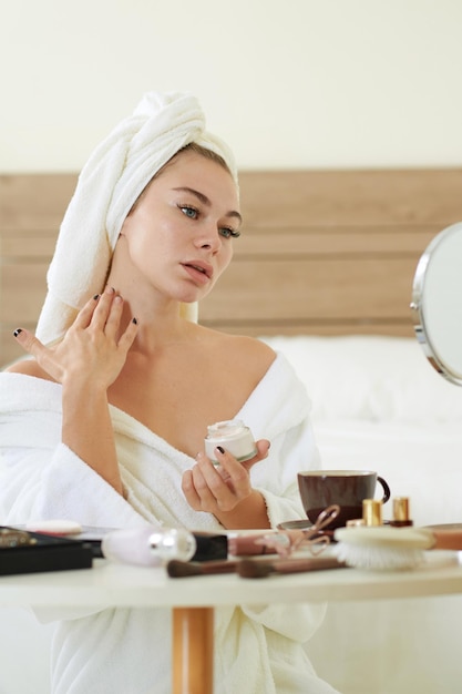 Young woman applying cream on neck and chest that helps to firm skin and reduce wrinkles