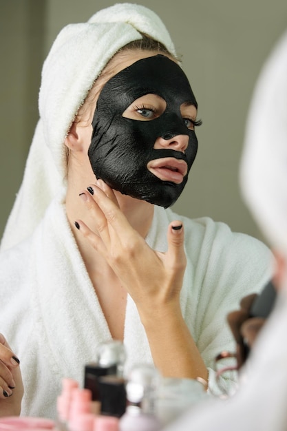 Photo young woman applying deep cleanse black sheet mask in front of bathroom mirror