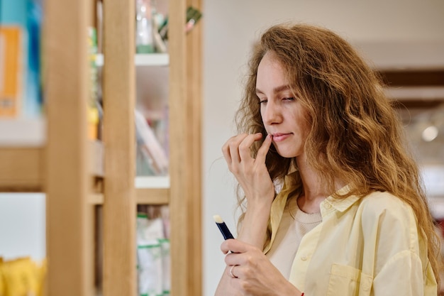 Young woman applying lipstick on her lips while doing shopping in cosmetics store