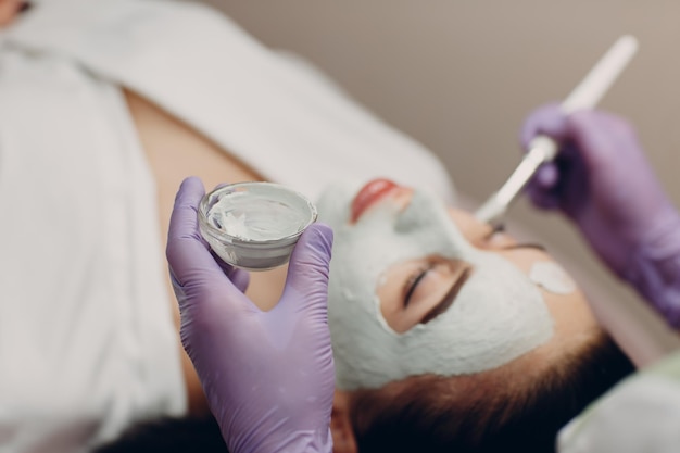 Young woman applying mask of clay on face in beauty spa