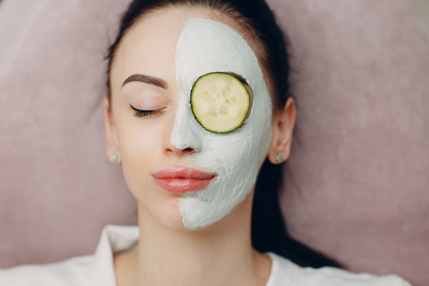 Young woman applying mask of clay on face with cucumber on eyes in beauty spa.