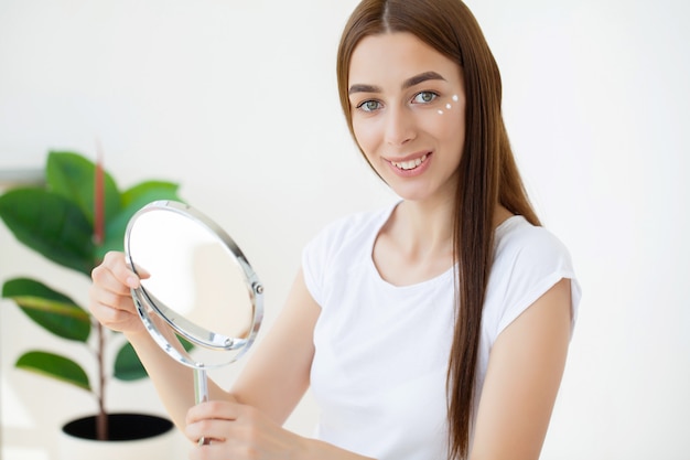 Young woman applying moisturizer on her face in front of mirror