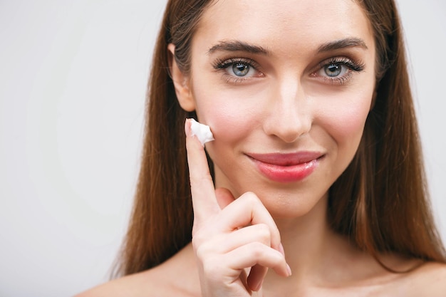 Photo young woman applying moisturizing lotion cosmetic cream on face for clean healthy soft skin care