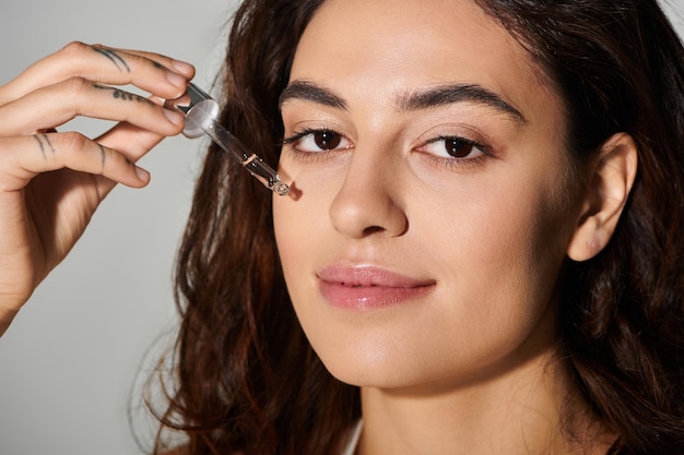Young woman applying serum to her face with a dropper in a neutral background setting
