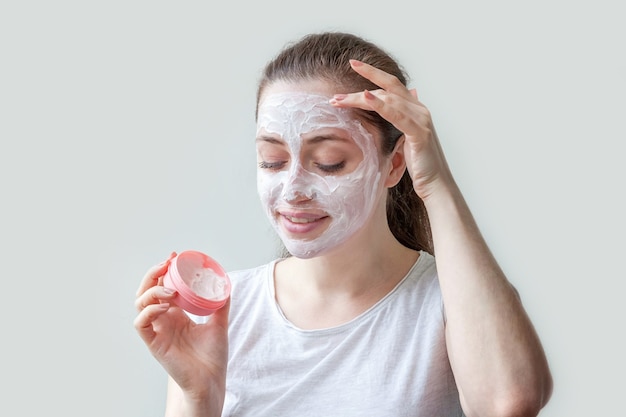 Young woman applying white nourishing mask or creme on face isolated on white