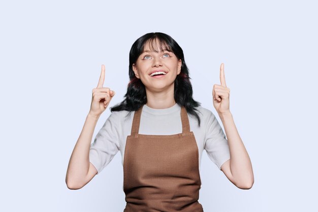 Photo young woman in apron showing hands up to empty space for your advertisement