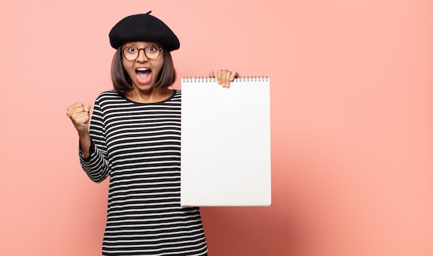 Young woman artist feeling shocked, excited and happy, laughing and celebrating success, saying wow!