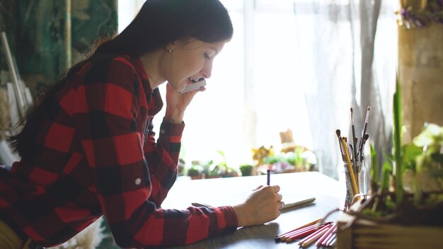 Молодая женщина-художник рисует эскиз на бумажном блокноте карандашом и разговаривает по телефону в помещении