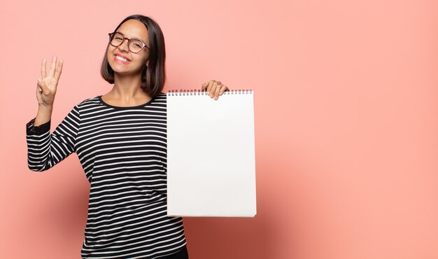 젊은 여성 아티스트가 미소하고 친절하게보고, 앞으로 손으로 세 번째 또는 세 번째를 보여주는, 카운트 다운