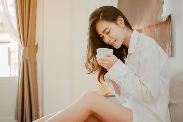 Young woman asia living at home relaxing and drinking cup of hot coffee in the bedroom on holiday. Asian, asia, relax, alone, technology, lifestyle concept.