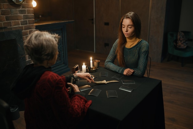 Young woman asking advice at mature old gypsy taro reader. Female client and fortune teller sitting at table with burning candle