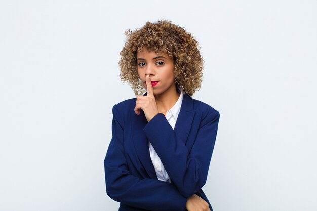 Young woman   asking for silence and quiet, gesturing with finger in front of mouth, saying shh or keeping a secret against  wall