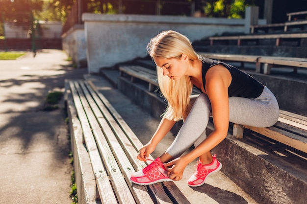 夏に運動場のベンチに座ってスニーカーを結ぶ若い女性アスリート。トレーニングの準備。いい結果