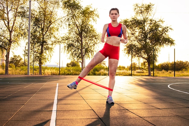 切断された腕を持ち、スポーツフィールドで体を燃やす若い女性アスリートレジスタンスバンドは日没時に屋外で運動します