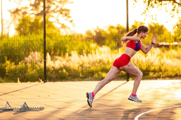 Young woman athlete with an amputated arm and burns on her body runs around the sports field She trains running from the starting block outdoors at sunset