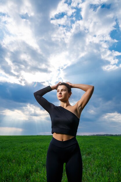 Young woman of athletic build with a black top and black leggings on a background of blue sky. Healthy lifestyle, athletic brunette girl. The concept and motivation of sports