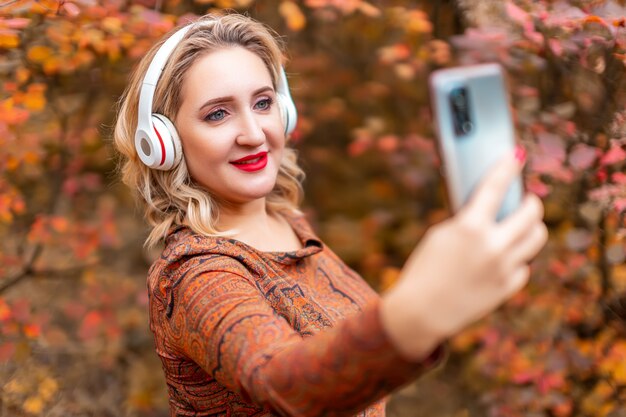 A young woman on the background of an autumn park takes a selfie on her phone