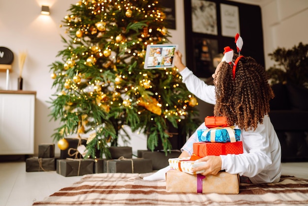 Photo a young woman on background of christmas tree with gifts with tablet has video call or video chat
