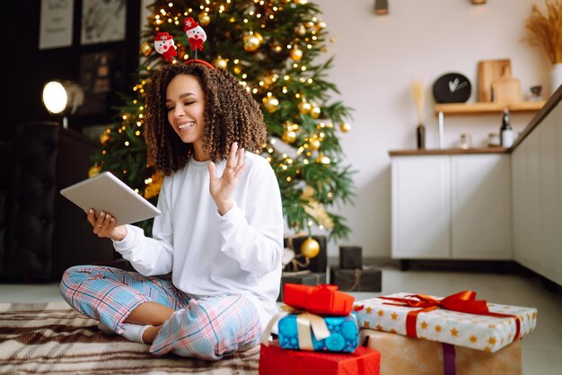 크리스마스 트리 배경에 태블릿을 들고 선물을 들고 있는 한 젊은 여성이 화상 통화나 화상 채팅을 합니다.